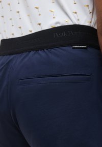 Gros plan d'une personne portant un pantalon bleu marine avec une ceinture élastique noire et une poche passepoilée à l'arrière, associé à une chemise blanche avec de petits motifs dorés.