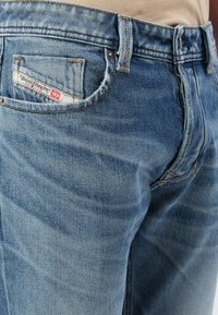 Des jeans en denim bleu clair présentent une texture usée avec des effets de frottement subtils, une étiquette sur la poche arrière et des coutures jaunes contrastantes.