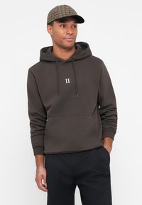 Sweatshirt à capuche marron avec poche avant, détail imprimé blanc "II," porté avec une casquette à carreaux. Le tissu est lisse et extensible.