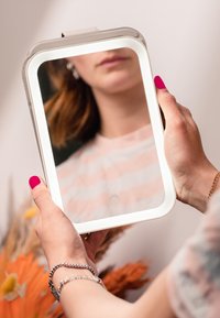 Beleuchteter rechteckiger Spiegel mit einem weißen LED-Rahmen, der von einer Hand mit rosa Nagellack gehalten wird. Im Hintergrund sind weich verschwommene Pflanzen zu sehen.