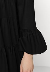 Vestido negro con una silueta fluida, tejido texturizado, niveles fruncidos y mangas de campana que acentúan el diseño. Estilo sencillo y versátil.