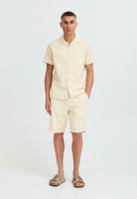 Lys beige kortermet skjorte med lomme, matchende shorts og brune sandaler. Myk tekstur, avslappet passform, brettede ermer på shortsene.