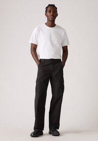 Witte katoenen T-shirt, relaxte pasvorm, zwarte cargobroek met meerdere zakken en zwarte gestructureerde sneakers, staand tegen een neutrale achtergrond.