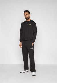 Conjunto de sudadera y pantalones negros, con un estampado de logo en amarillo y blanco. Tela suave, corte relajado, con cinturilla elástica y bolsillos laterales.