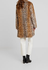 Abrigo largo de estampado de leopardo en pelo sintético en tonos beige y crema, con corte recto y mangas largas, combinado con pantalones blancos y botas negras hasta el tobillo.