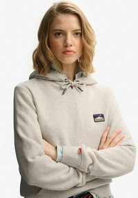 Felpa con cappuccio grigio chiaro con coulisse, logo frontale a toppa e polsini a costine indossata sopra una camicia a righe. Tessuto liscio e vestibilità rilassata.