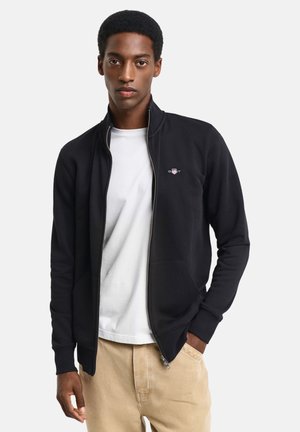 Schwarzer Zip-Up-Hoodie mit hohem Kragen, Seiten Taschen und Logo. Über einem weißen Shirt getragen; das Model steht mit den Händen in den Taschen.
