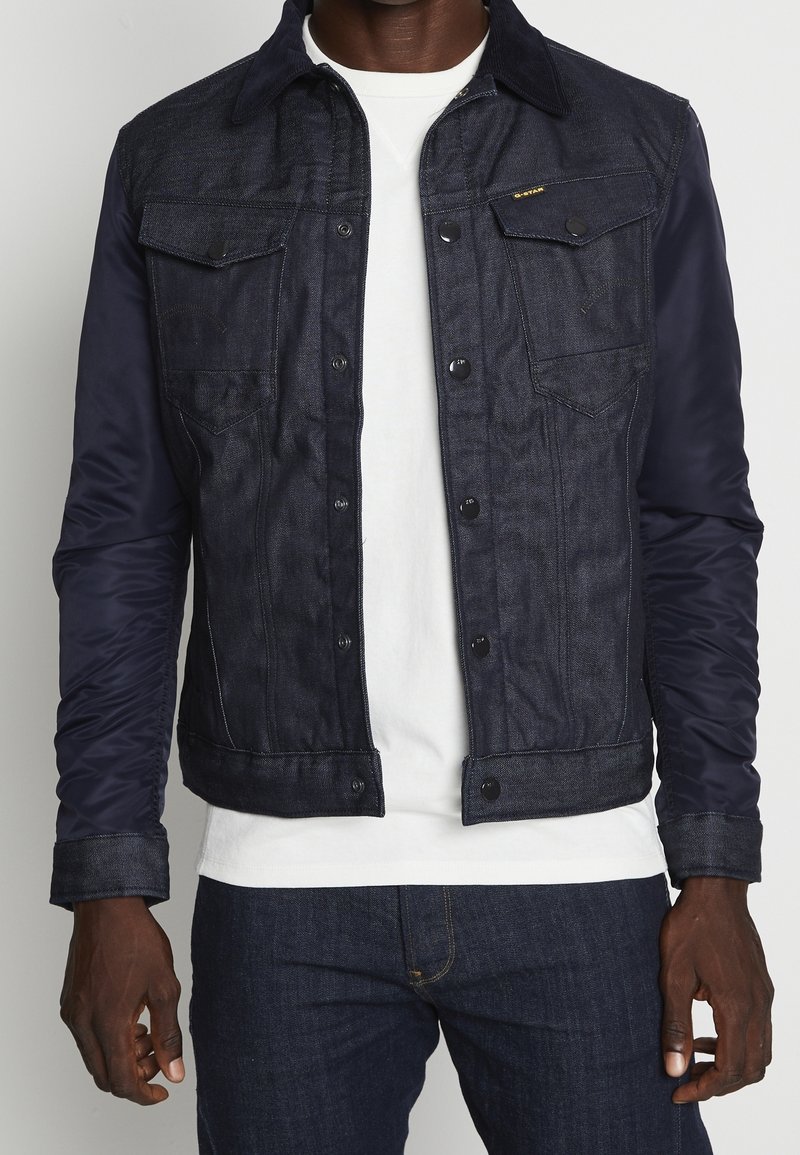 Mörk denimjacka med marinblå vadderad tyg på ärmarna. Har knäppning med knappar, två bröstfickor och kontrasterande sömmar längs med sömmarna.