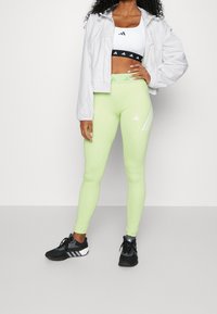 Femme portant des leggings taille haute vert clair, un soutien-gorge de sport blanc, une veste blanche et des chaussures de sport noires, debout contre un mur uni.