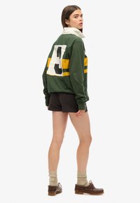 Sweatshirt vert avec des rayures jaunes et un grand numéro blanc dans le dos, associé à un short foncé et des chaussures marron, avec des chaussettes texturées.