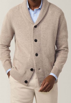 Cardigan a maglia beige con collo a scialle, dotato di bottoni scuri e polsini a coste. Indossato sopra una camicia a righe blu chiaro.