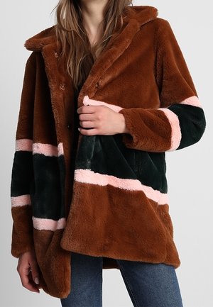 Kvinde iført en brun faux pelsjakke med sorte og pink vandrette striber, delvist knappet, kombineret med blå jeans.