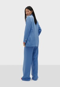 Set blu composto da un blazer su misura e pantaloni a gamba larga. Il blazer presenta dettagli con bottoni sulle maniche. Tessuto liscio con un design strutturato.