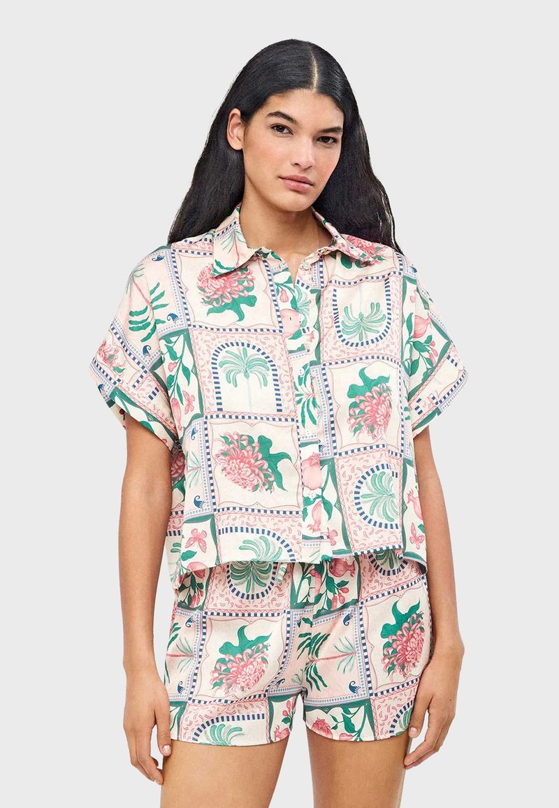 Femme aux longs cheveux bruns portant une chemise à manches courtes à boutons et un short assortis à motif floral, sur un fond uni.