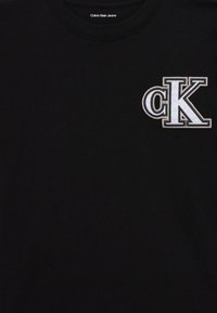 Fekete kerek nyakú póló, bal mellen fehér hímzett "cK" logóval, a gallér belsejében pedig "Calvin Klein Jeans" felirattal.