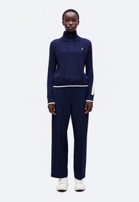 Femme debout portant un pull en maille torsadée bleu marine, un pantalon large assorti et des baskets blanches sur un fond blanc uni.