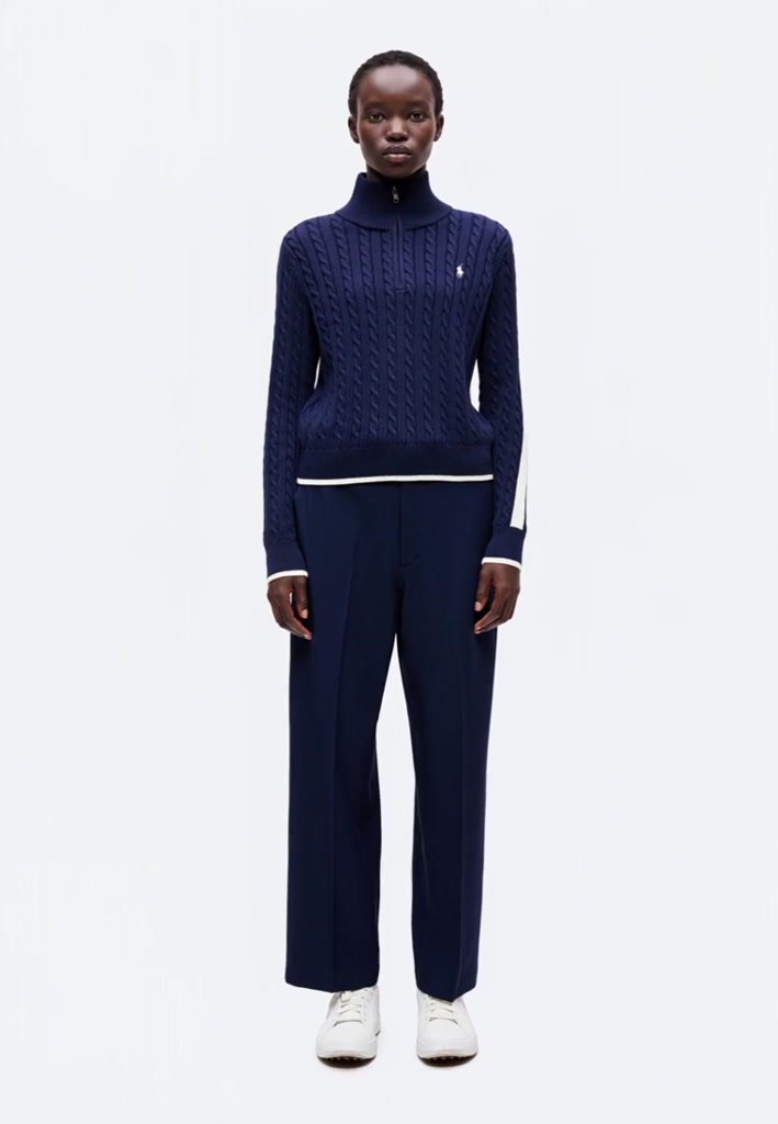 Femme debout portant un pull en maille torsadée bleu marine, un pantalon large assorti et des baskets blanches sur un fond blanc uni.