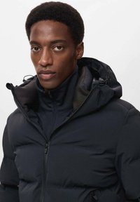 Giacca a vento in blu scuro con zip anteriore e cappuccio. Presenta sezioni imbottite per isolamento e una finitura liscia. Inclusi taschini con zip.