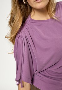 Lilac blouse with soft, draped fabric; features gathered detailing at the shoulders and short sleeves. Smooth texture and rounded neckline.