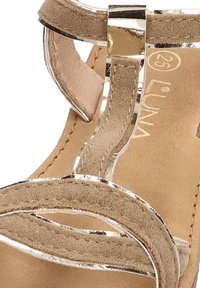 Sandalia de ante beige con correas metálicas, tacón plano y un forro interior suave. Destaca por su diseño de correas cruzadas y acentos brillantes.