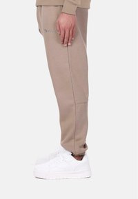 Pantalon de jogging beige avec une taille côtelée, des poches latérales et un texte brodé. Associé à des baskets blanches avec une semelle épaisse.