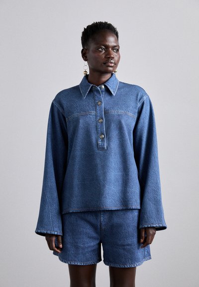 Haut en denim avec un col, une patte de boutonnage et des manches longues, associé à un short en denim. Les deux pièces sont dans une teinte de bleu moyen.