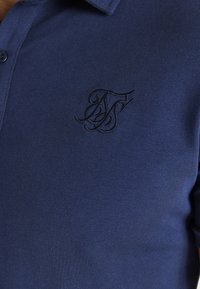 Paita polo tummansinisenä, valmistettu sileästä puuvillakankaasta, jossa on kaulus ja rintaa koristaa tummansinisellä brodeerattu logo.