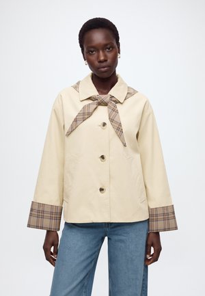 Femme portant une veste beige avec col, poignets et détail noué à l'avant à carreaux, associée à un jean bleu, debout devant un fond blanc uni.