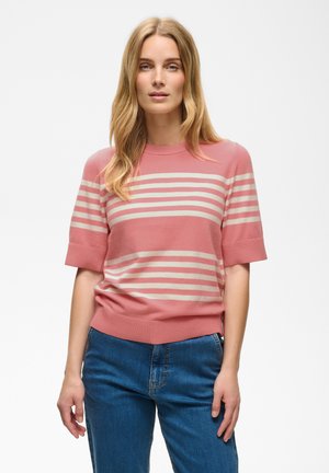 Femme aux longs cheveux blonds portant un pull rose à manches courtes avec des rayures blanches et un jean bleu, debout devant un fond clair et uni.