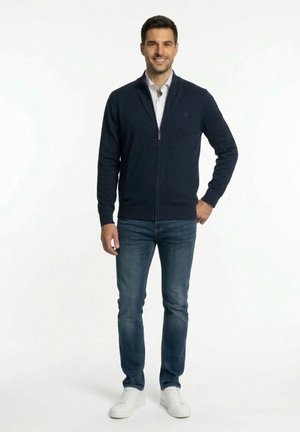 Hombre sonriendo, vestido con suéter azul marino con cremallera, camisa de cuello gris claro, jeans azules y zapatillas blancas, de pie contra un fondo blanco liso.