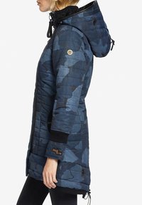 Kvinde set fra siden iført en blå og sort camouflage-mønstret quiltet hættejakke med lynlås og strikkede manchetter.