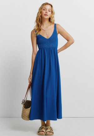 Vrouw draagt een mouwloze blauwe maxi-jurk met gehaakt lijfje, houdt een geweven strooien tas vast en draagt beige sandalen, staat tegen een witte achtergrond.