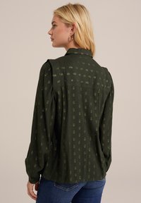 Blusa verde scuro con un tessuto testurizzato, caratterizzata da piccoli motivi a pois dorati, colletto e maniche lunghe. Finitura con una vestibilità leggermente morbida.