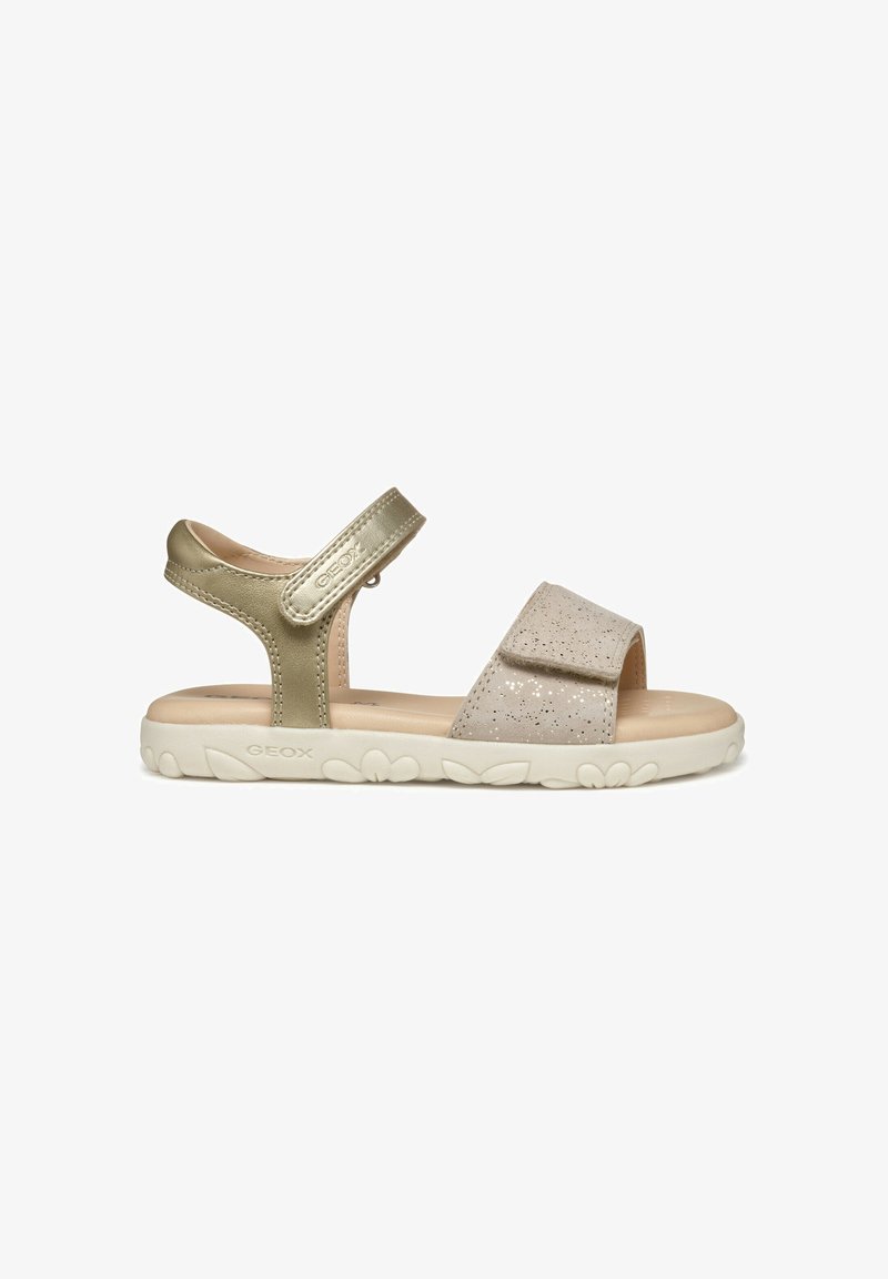 Gouden en beige sandalen met een gestructureerde band en een glinsterende bovenkant. Klittenbandsluiting en een gewatteerde witte zool. Geox-merk zichtbaar.