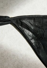 Schwarze transparente Dessous mit filigranen Spitzenmustern und doppelten Taillenbandriemen, flach auf einer hell strukturierten Oberfläche ausgelegt.
