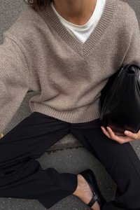 Bēšas adītas džempera ar V veida kakla izgriezumu, savienotas ar melnām platām biksem. Melna rokassoma turēta vienā rokā. Gluda, teksturēta auduma.