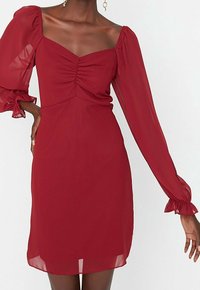 Rotes Kleid mit quadratischem Ausschnitt, gerafftem Vorderteil und transparenten Puffärmeln. Weicher Stoff, knielange Länge und Details mit gefütterten Ärmeln.