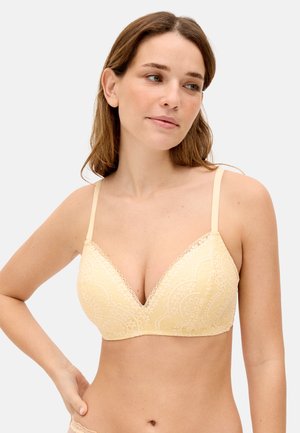 Mujer con cabello castaño claro que lleva un sujetador de encaje amarillo pálido, mirando ligeramente hacia su izquierda con una expresión neutral.