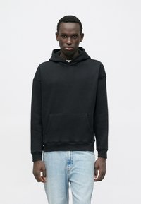 Felpa nera con cappuccio in tessuto morbido, con tasca frontale e polsini a costine, abbinata a jeans in denim azzurro chiaro.