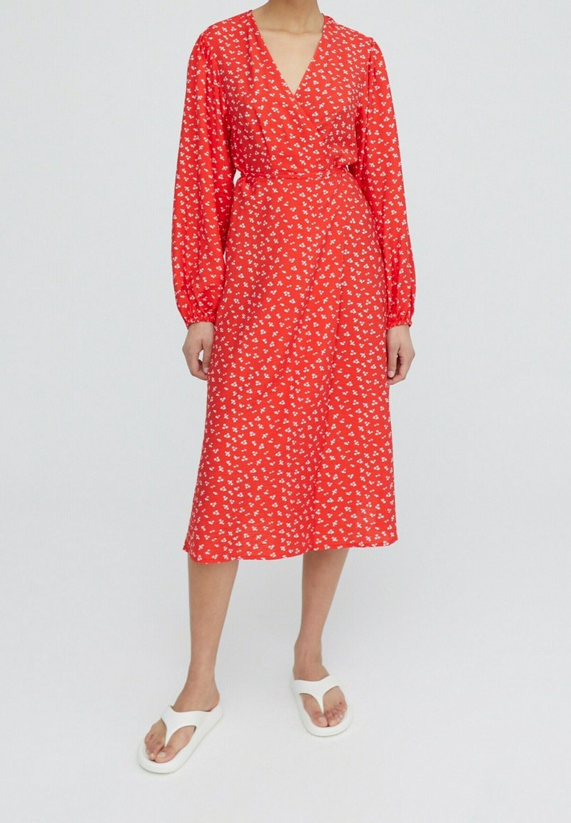 Vestido midi rojo con diseño envolvente, mangas largas de globo y patrones florales blancos. Llevado con chanclas blancas. Textura de tela suave.