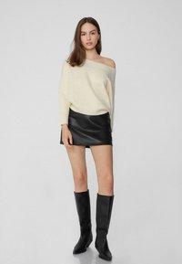 Off-white gestrickter Pullover mit weitem Ausschnitt, kombiniert mit einem schwarzen Leder-Minirock und knielangen schwarzen Stiefeln mit Absatz. Schlichte Gestaltung, strukturiertes Material.