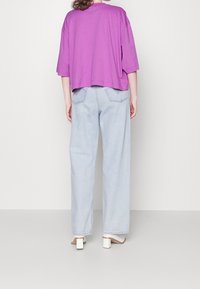 Camiseta morada de gran tamaño con mangas cortas, combinada con pantalones de mezclilla de tiro alto y patas anchas en azul claro y sandalias blancas de tacón blok.