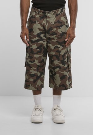 Mand iført knælange camouflage cargo shorts, hvide sokker, hvide sneakers og en sort skjorte mod en ensfarvet grå baggrund.