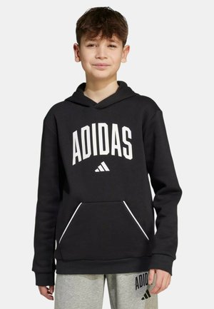 Garçon portant un sweat à capuche Adidas noir avec logo blanc et un pantalon de survêtement Adidas gris, debout devant un fond clair uni.