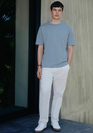 Junger Mann steht mit einer Hand in der Tasche, trägt ein blau gestreiftes T-Shirt, weiße Hose, weiße Turnschuhe und ein Perlenarmband drinnen.