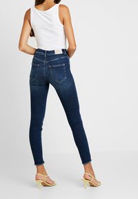 Jeans de mezclilla de color azul oscuro con cintura alta, ajuste slim y dobladillo deshilachado. Los bolsillos traseros tienen una forma rectangular estándar. Combinados con tacones amarillos con tiras.