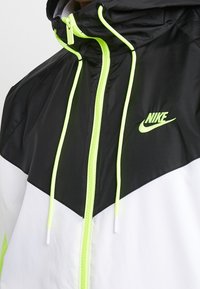 Nike-jacka med en svart överdel, vit underdel och neongröna detaljer. Inkluderar en dragkedjefram och justerbar huva.