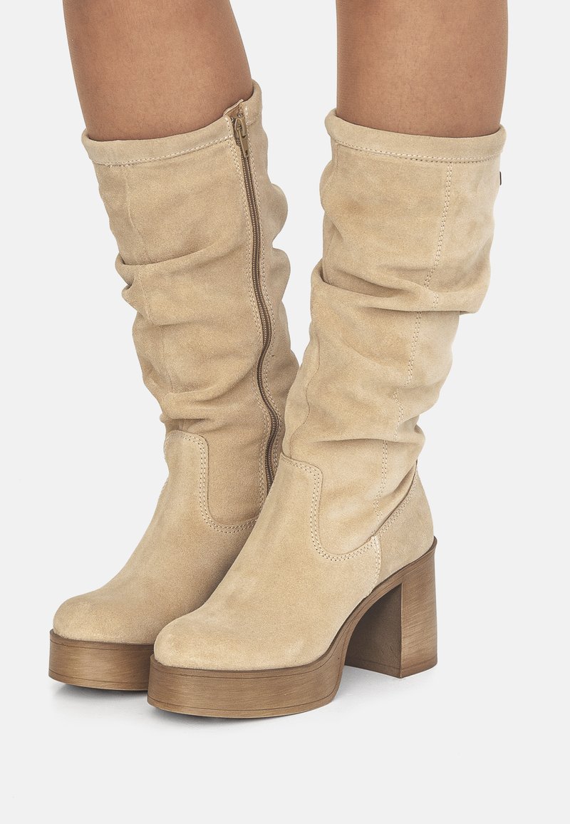 Stivali al ginocchio in suede beige con design slouchy, chiusura con zip laterale e un tacco spesso in legno. Caratterizzati da dettagli cuciti e una texture liscia.