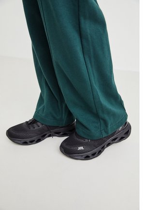 Baskets noires de sport avec des semelles en maille et en caoutchouc, présentant un design distinctif avec des découpes et un logo. Assorties à un pantalon de survêtement teal.