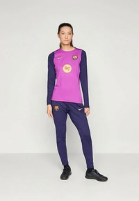 Woman standing wearing purple and navy football tracksuit with Nike and FC Barcelona logos, black cleats, against a plain light background.